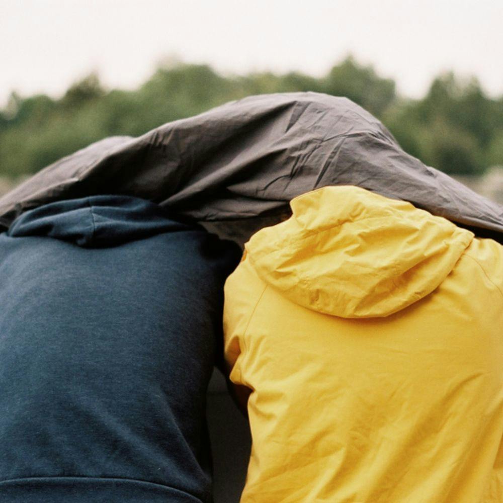 zwei aneinander lehnende Personen sind von hinten zu erkenne, sie schützen sich mit Jacken vor dem Regen