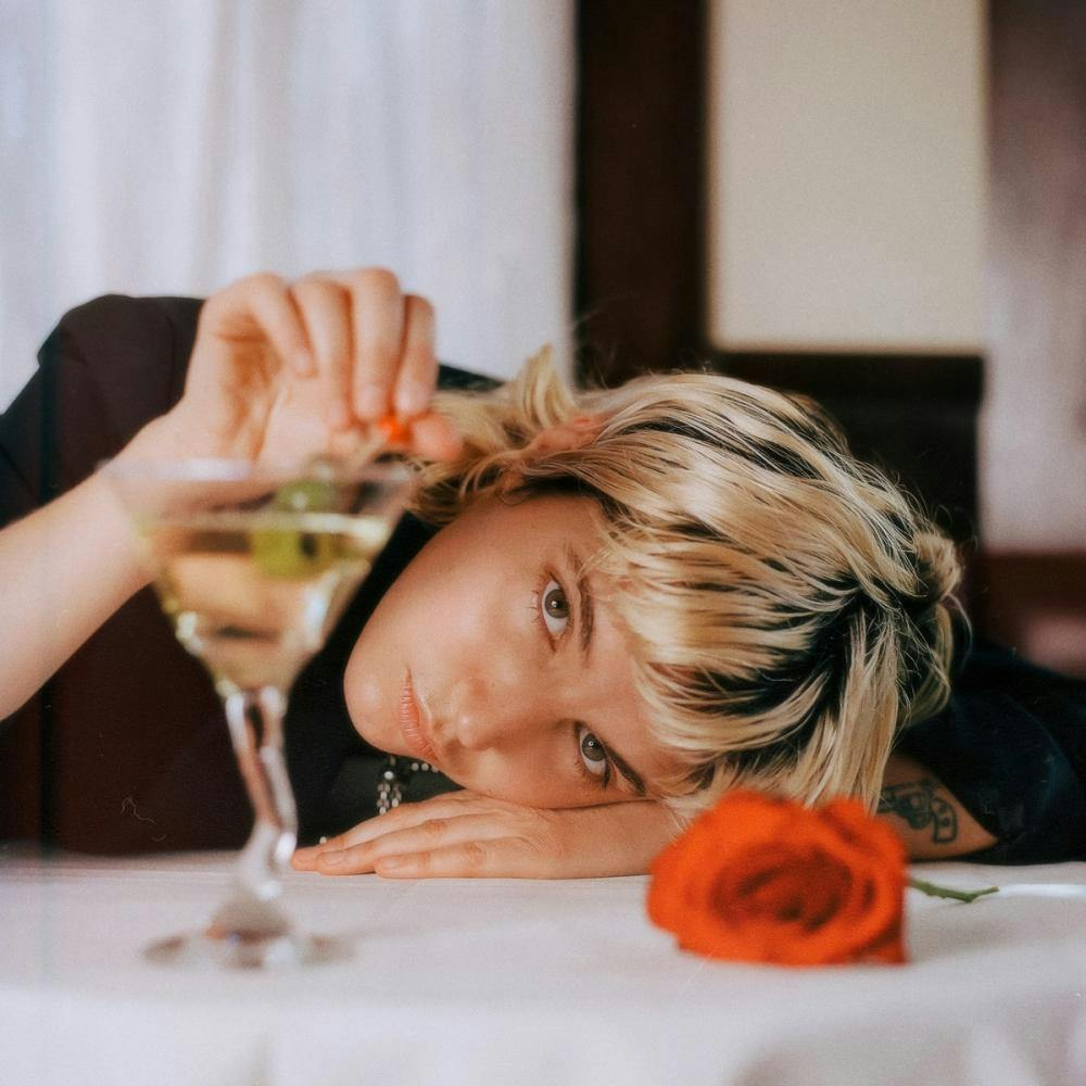 Die Künstlerin Caroline Rose liegt mit ihrem Kopf auf einem Tisch, ihre Hand auf einem Glas und vor ihr eine Rose. 
