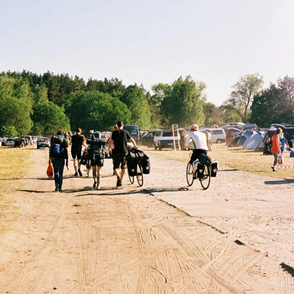 Viele Leute laufen mit ihren Rädern Richtung Festivaleingang, rechts neben ihnen sind Zelte und links neben ihnen parkende Autos.