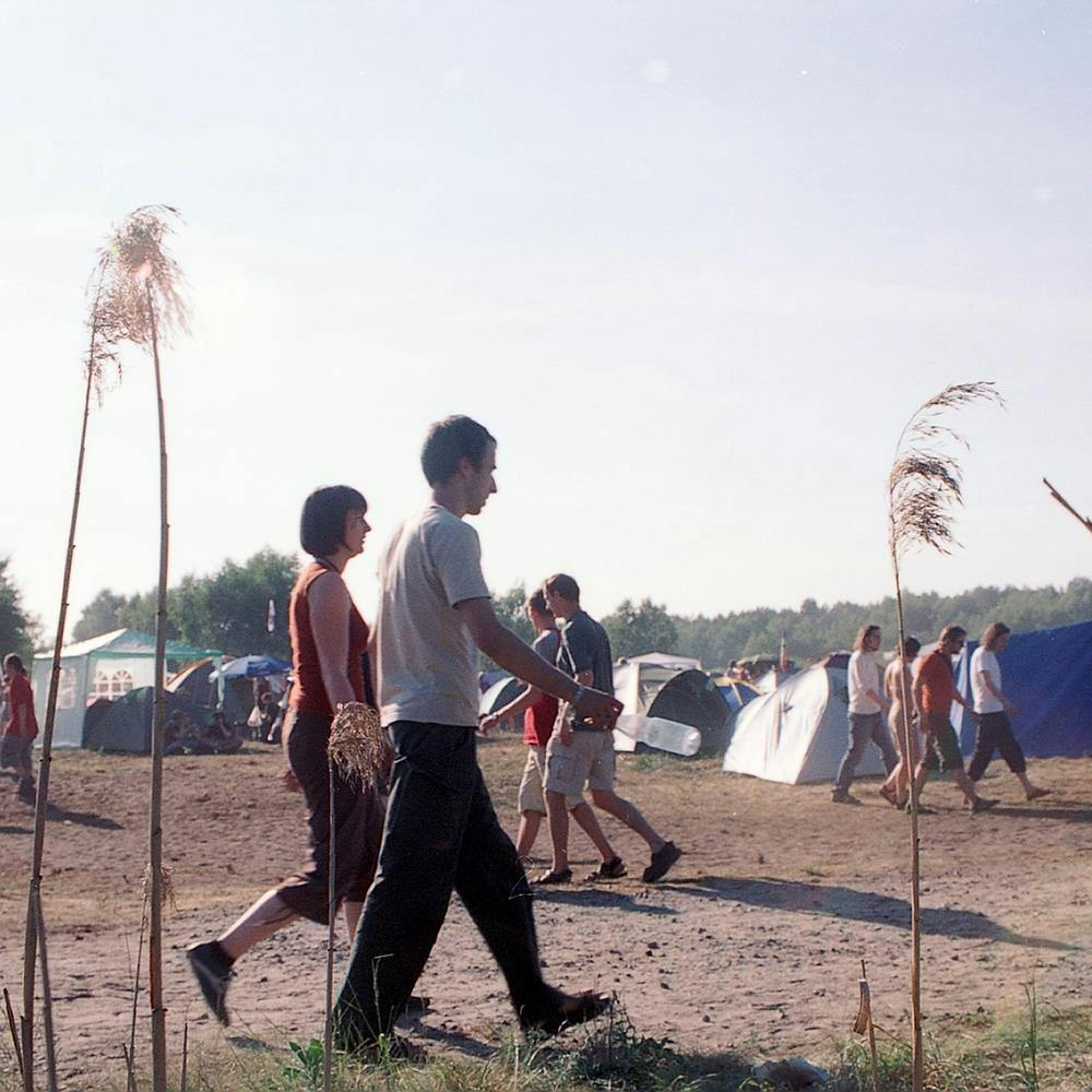 Ein Mann und eine Frau laufen über den Zeltplatz, es ist heiß, die Sonne scheint, im Hintergrund sind Zelte zu sehen, im Vordergrund des Bildes sind einzelne Grashalme.